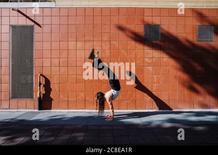 Giovane donna che esegue handstand sul marciapiede contro parete piastrellata in città Foto Stock