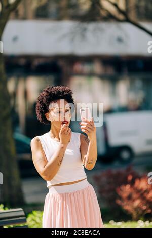 Donna mid adulta che applica il rossetto mentre si guarda nello smartphone Foto Stock