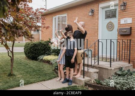 La famiglia indossa maschere agitando le mani mentre si trova fuori casa dentro iarda Foto Stock