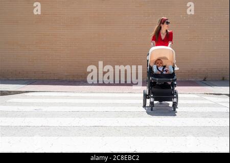 Madre che indossa occhiali da sole che spinge il figlio nel carrello del bambino mentre cammina sulla strada Foto Stock