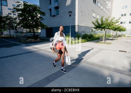 Giovane donna con borsa per lo shopping e skateboard a piedi sul sentiero in città Foto Stock