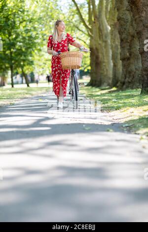 Donna che guarda via mentre cammina con la bicicletta sul sentiero dentro parco pubblico durante il giorno di sole Foto Stock