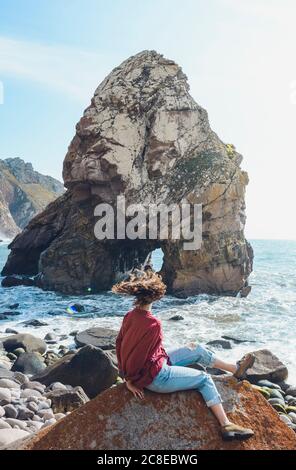 Donna spensierata scuotendo la testa mentre si siede sulla roccia a Ursa Beach, Portogallo Foto Stock