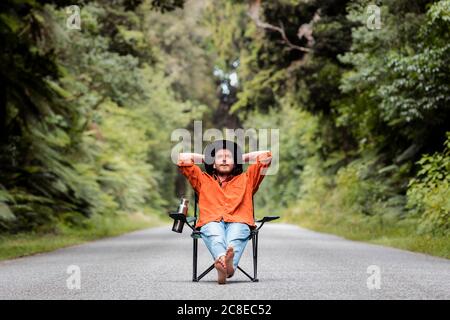 Uomo premuroso con le mani dietro la testa seduta in sedia a. su strada di campagna tra alberi Foto Stock