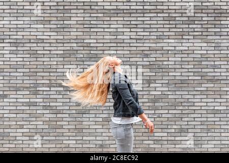 Donna che lancia i capelli lunghi mentre si sta in piedi da parete grigia del mattone Foto Stock