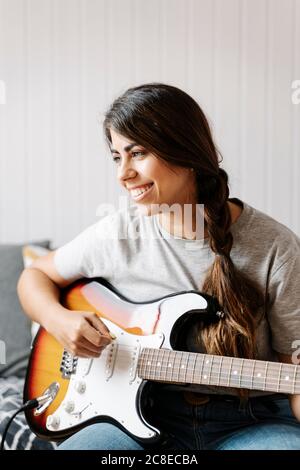 Donna felice che suona la chitarra elettrica mentre si siede a casa Foto Stock