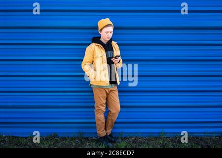 Ragazzo che usa lo smartphone mentre si sta in piedi contro l'otturatore blu Foto Stock