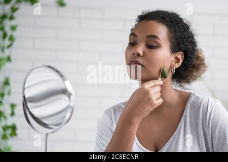 Primo piano di giovane donna massaggiando il viso con rullo di giada mentre guardando nello specchio a casa