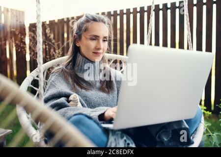Bella giovane donna che usa il computer portatile mentre si siede in swing in giardino Foto Stock