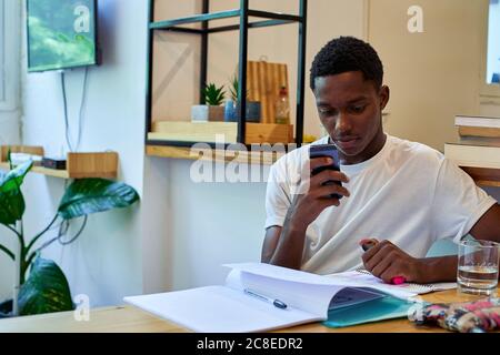 Giovane uomo che usa lo smartphone mentre studia sul tavolo a. casa Foto Stock