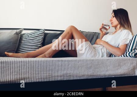 Donna che si rilassa a casa accogliente in soggiorno seduta sul divano bere acqua. Routine mattutina Foto Stock