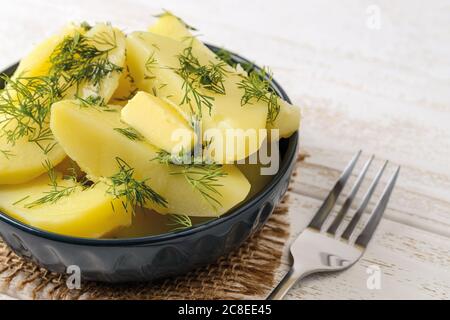 Primo piano di patate appena bollite con burro e aneto in una ciotola di ceramica e forchetta su un tavolo di legno bianco. Cuocete a casa. Foto Stock