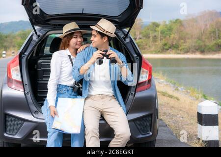 Giovane coppia asiatica con uomo turista che tiene binocoli con ragazza sorridente seduto vicino con mappa su tronco auto in vista campo rurale con lago e m Foto Stock