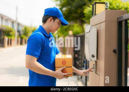 Consegna manasiatica giovane uomo in uniforme blu con scatola di pacchi sorriso e squillo clienti campana porta in casa villaggio con spazio copia. Pubblicità Foto Stock