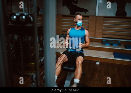 Giovane uomo muscolare fitness con maschera medica facendo esercizio pesante di peso per schiena e braccio sulla macchina in palestra.prevenzione di COVID - 19 coronavirus Foto Stock
