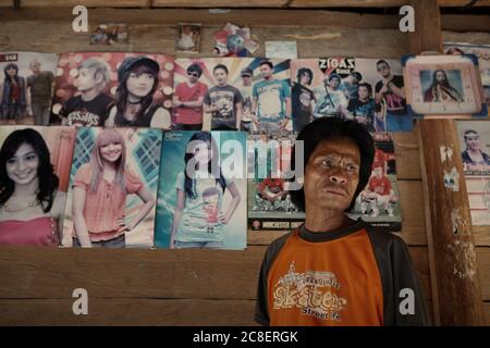 Ritratto di Umbu Tiu, un uomo di famiglia Sumbanese fuori dalla sua casa tradizionale a Tebara, villaggio di Praijing, Isola di Sumba, Indonesia. La sua casa fu costruita interamente con materiali provenienti dalla natura. Vecchie immagini di artisti stranieri e locali e una foto della squadra del Manchester United sulla parete esterna (sfondo) sono tra le decorazioni molto limitate di origine moderna della casa. Foto Stock