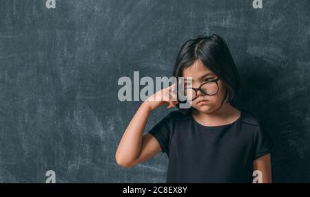 Ritorno a scuola. Ragazza piccola seria in occhiali pensando di toccare la testa con un dito vicino a lavagna. Bambino della scuola elementare. Foto Stock