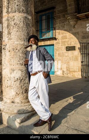 Havana / Cuba - 04.16.2015: Afro cubano vecchio con una lunga barba bianca che indossa un vestito e una berta militare nera, fumando un grande sigaro Foto Stock