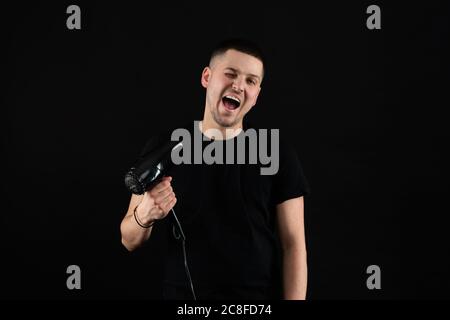 Divertente giovane uomo 20s anni in nero t-shirt tenere soffio capelli asciutti isolato su sfondo nero parete, studio ritratto Foto Stock