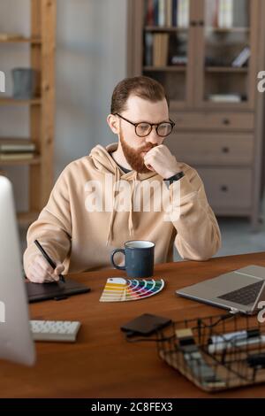 Pensieroso giovane designer in felpa con cappuccio seduto a tavola di legno e analizzare la tavolozza dei colori per il web design Foto Stock