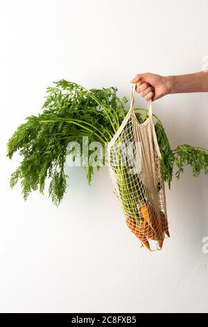 Mano maschile con carota in una griglia di cibo su uno sfondo bianco parete, un agricoltore tiene verdure appena raccolte con foglie verdi, raccolto fresco Foto Stock