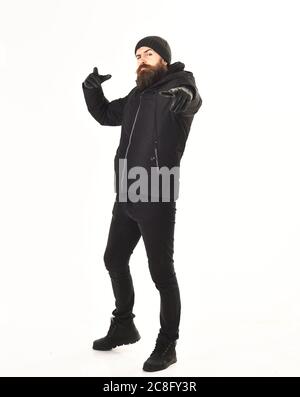 Hipster con il viso stretto indossa abiti caldi e punta in avanti. Ragazzo elegante in giacca e cappello, isolato su sfondo bianco. L'uomo con barba indossa un abito invernale nero e brutale. Concetto di outfit invernale. Foto Stock