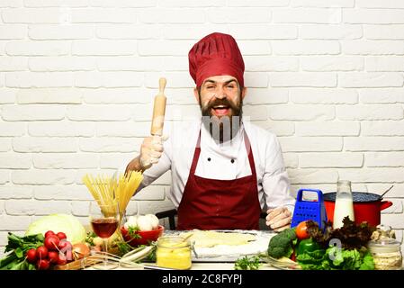 Concetto di cucina professionale. L'uomo con la barba tiene il perno su sfondo bianco in mattoni. Cuoco con il viso allegro in uniforme di borgogna siede a tavola con verdure e utensili da cucina. Chef prepara l'impasto Foto Stock
