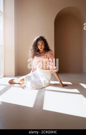 Vista frontale del meraviglioso modello di giovane brunetta semi-africana con outfit estivo alla moda guardando la macchina fotografica. Mulatto ragazza con lunghi capelli neri ricci che si posano sotto la luce del sole sul pavimento vicino alla grande finestra. Foto Stock