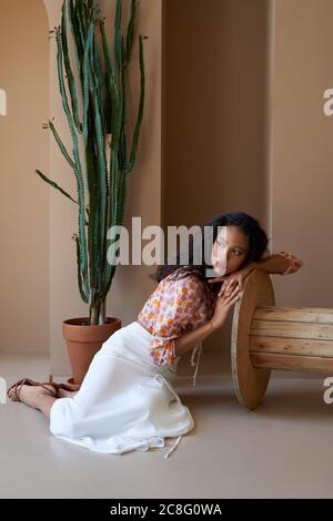 Bellissima ragazza mulatto con capelli ricci che posano vicino a pianta succulente alta in vaso di fiori, seduta sul pavimento e appoggiata su ruota di legno. Splendida donna semi africana che indossa blusa, gonna bianca e tacchi. Foto Stock