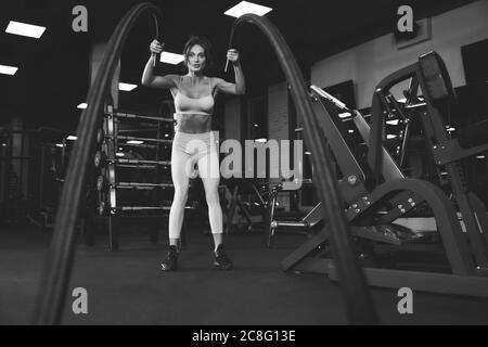 Dal basso vista di donna brunette in leggero sport allenamento in palestra vuota con corde. Ritratto monocromatico a lunghezza intera di attraente donna in forma con il viso forte che fa cardio. Concetto di sport. Foto Stock