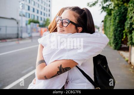 Due donne con braccia tatuate in piedi sul marciapiede, abbracciando. Foto Stock