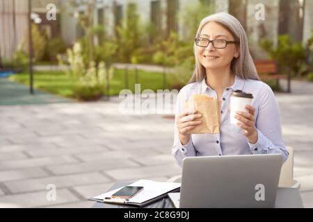 Lavorare online. Ritratto di una donna d'affari felice matura che indossa occhiali con pranzo, caffè e panino mentre lavora all'aperto, utilizzando un computer portatile. Uomini d'affari, freelance Foto Stock