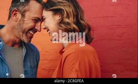 Bella coppia innamorata che tocca le fronte e sorridente. Coppia felice in piedi contro parete di colore rosso e arancione. Foto Stock
