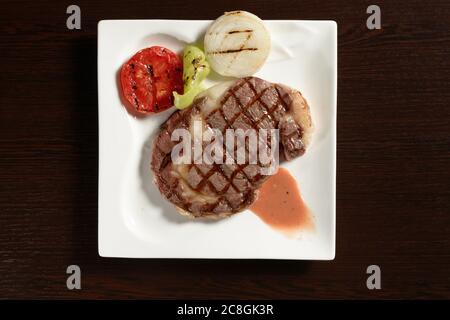 Bistecche di manzo in marmo fritte con verdure grigliate di pomodoro, cipolla e peperone su un piatto quadrato su un tavolo di legno. Vista dall'alto. Foto per ristorante Foto Stock