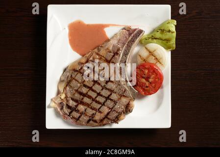 Bistecca di manzo cowboy alla griglia con verdure grigliate di pomodoro, cipolla e peperone su un piatto quadrato su un tavolo di legno. Vista dall'alto. Foto per ristoratore Foto Stock