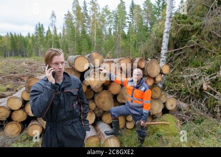 Uomo maturo che controlla legno tritato con giovane uomo che parla sul telefono cellulare fuori della foresta Foto Stock