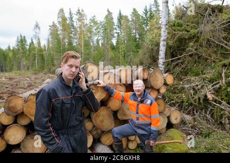 Uomo maturo che controlla legno tritato con il giovane uomo usando il telefono fuori della foresta Foto Stock