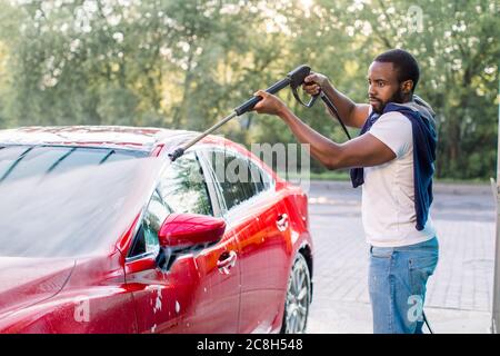 African elegante giovane uomo messo a fuoco in vestiti casual che lavano l'auto rossa con una pistola ad acqua sulla stazione self-service di lavaggio auto all'aperto, sciacquando il Foto Stock