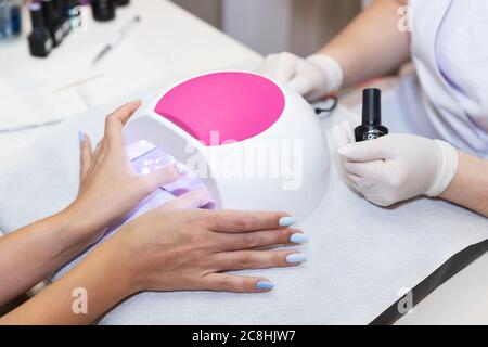 Primo piano bella donna mani e lampada per unghie sul tavolo. Salone gel unghie. Lampada UV. Processo di essiccazione del gel sulle unghie. Bellezza e cura di sé. Tecnologia moderna Foto Stock