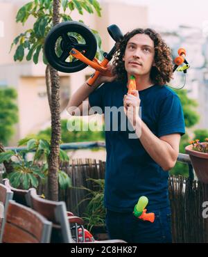 Il giovane uomo del giorno del Padre con pistole ad acqua e una bicicletta in mano, stanco dopo aver giocato con i bambini, si erge sul balcone in una T-shirt blu e je Foto Stock