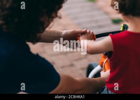 Festa del Padre. Papà con il figlio piccolo su una bicicletta dal retro. Passeggiata in famiglia con un bambino. Attività all'aperto per famiglie. Papà aiuta il figlio con una bicicletta. Mani o Foto Stock