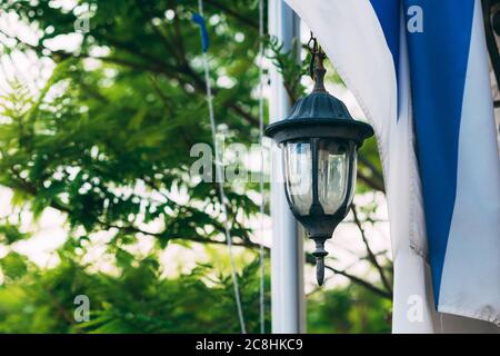 Bandiera israeliana e lampada da strada su uno sfondo di alberi verdi sfocati. Celebrazione della Giornata dell'Indipendenza di Israele. Giorno della memoria dell'Olocausto. Memorial Day fo Foto Stock