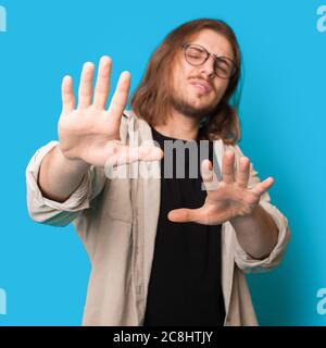 Uomo caucasico dai capelli lunghi con barba e occhiali che rifiuta qualcosa su una parete blu dello studio Foto Stock