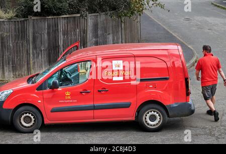 Un postino torna al suo luminoso furgone rosso Royal Mail (con il suo stemma reale) dopo aver postato un pacchetto in una casa. Foto Stock