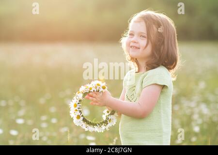 Sorridente bambina 2-3 anni facendo corona di fiori che si pone in campo sopra closeup di fondo soleggiato. Stagione estiva. Infanzia. Foto Stock