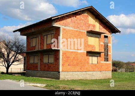 Blocchi da costruzione rossi abbandonati incompleti casa suburbana familiare senza facciata e con finestre parzialmente a bordo accanto alla strada lastricata Foto Stock