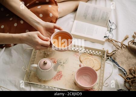 Una donna tiene una tazza di tè caldo sul letto a casa e legge un libro, tempo libero domestico Foto Stock