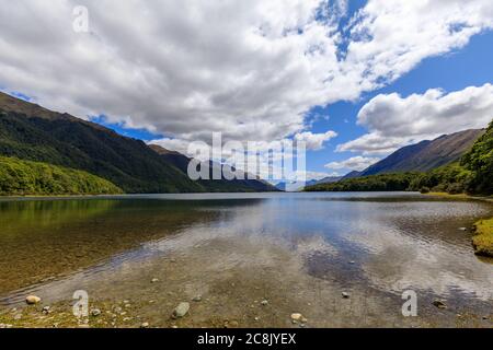 Le acque calme del Lago di Mavora Sud con montagne boscose lungo ogni lato e montagne in lontananza. Nuvole bianche e soffici su un cielo blu. Foto Stock