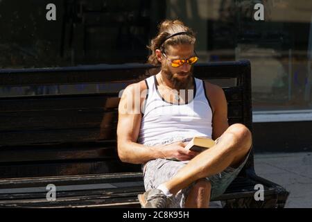 Durante la lettura di un libro, un uomo sedette su una panca di strada indossando occhiali da sole gialli e arancioni riflettenti, York, North Yorkshire, Inghilterra, Regno Unito. Foto Stock