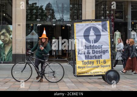 Glasgow, Scozia, Regno Unito. 25 luglio 2020. Una ragazza su Buchanan Street che indossa un cappello a cono del traffico con una bicicletta che tira un segno mobile che fa pubblicità al mercato dell'arte e del design all'aperto ora aperto in Royal Exchange Square. Credit: Notizie dal vivo SKULLY/Alamy Foto Stock